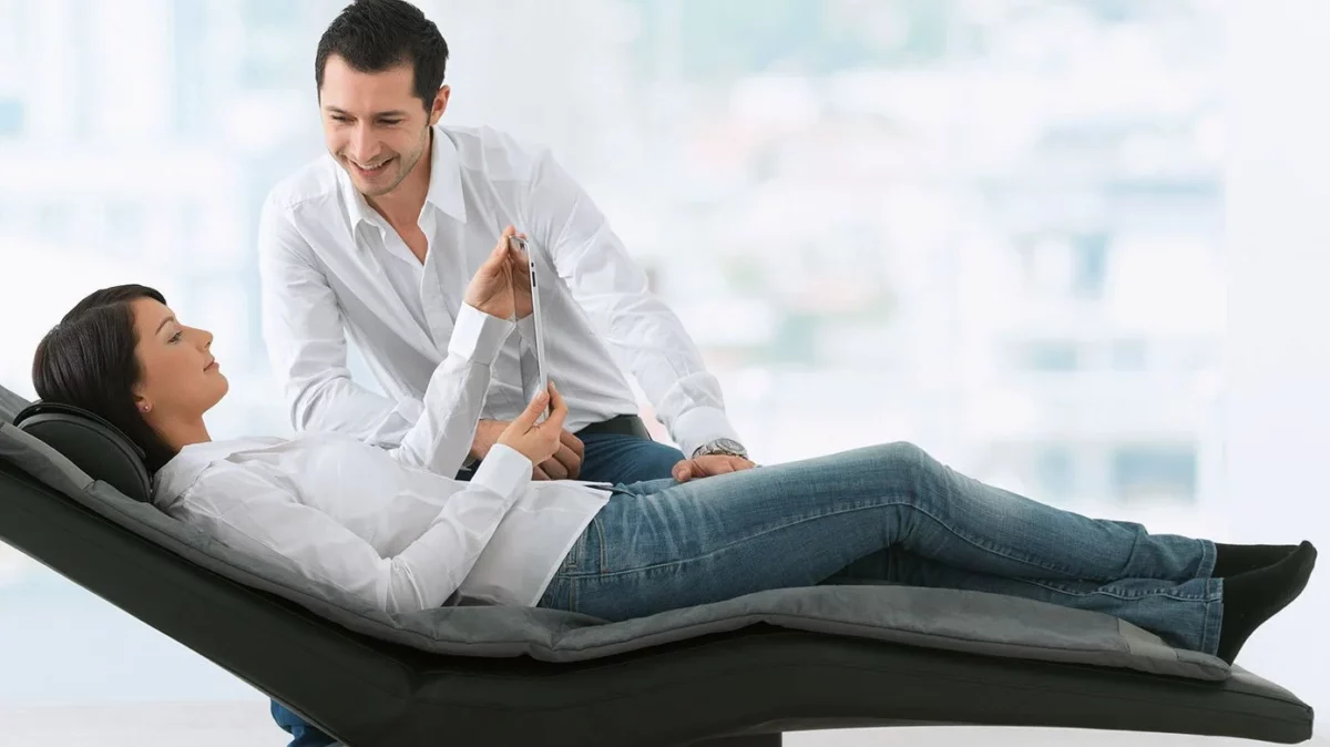 Frau liegt mit einem Tablet in der Hand auf einer verstellbarer Liege, Mann sitzt lächelnd daneben.