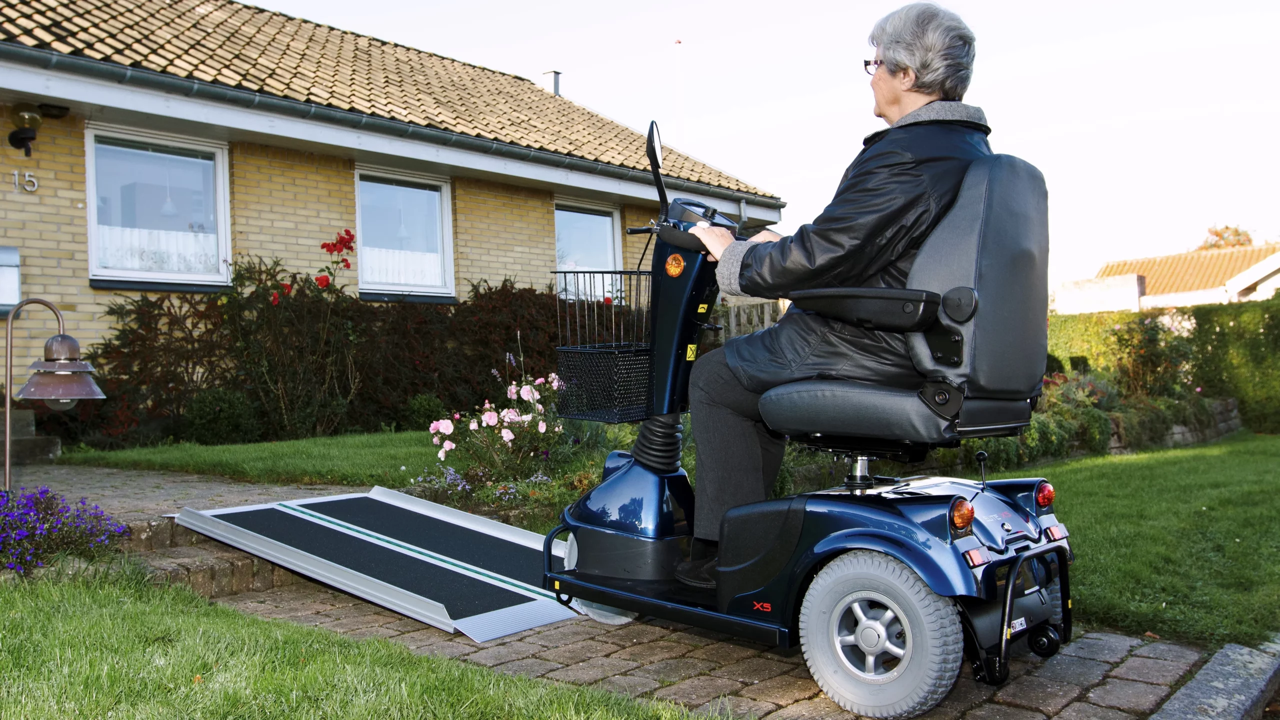 Ältere Frau fährt mit Elektromobil über Rampe zum Haus, zeigt Barrierefreiheit.