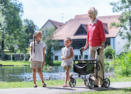 Ältere Frau geht mit Rollator spazieren, begleitet von zwei Kindern an einem Teich.