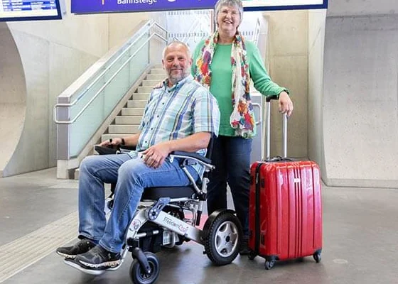 Paar am Bahnhof: Mann im Elektrorollstuhl, Frau steht mit Koffer neben ihm.