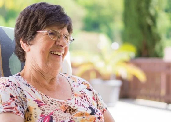 Ältere Frau sitzt entspannt im Freien und lächelt, freundliche Alltagsszene im Garten.