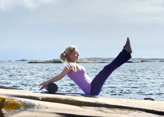 Frau macht Pilatesübung am Meer mit gestreckten Beinen und Pilatesrolle.